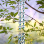 Pale green African Krobo bead wind chime with aqua crystal beads and fused glass pieces with raised glass bubbles, hanging outdoors among green leaves.