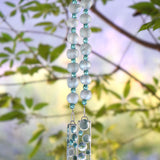 Pale green African Krobo bead wind chime with aqua crystal beads and fused glass pieces with raised glass bubbles, hanging outdoors among green leaves.
