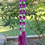Two strands of beaded wire hanging from a tree, anchored by two pieces of cranberry pink fused glass. Each strand has large, faceted acrylic beads in purple separated by smaller clear glass beads.