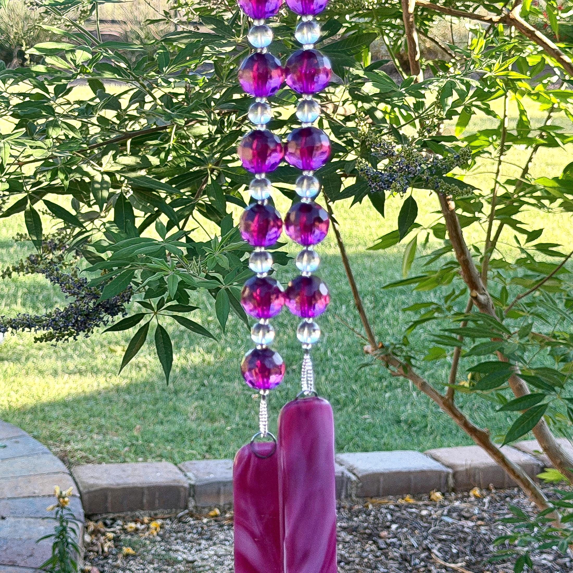 Two strands of beaded wire hanging from a tree, anchored by two pieces of cranberry pink fused glass. Each strand has large, faceted acrylic beads in purple separated by smaller clear glass beads.