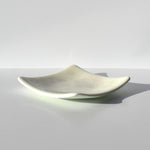A square glass dish with curved, upturned edges and a smooth, off-white surface. The center of the dish features a subtle, faint floral pattern in a light gray tone, integrated into the translucent material of the glass.