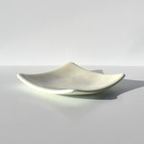 A square glass dish with curved, upturned edges and a smooth, off-white surface. The center of the dish features a subtle, faint floral pattern in a light gray tone, integrated into the translucent material of the glass.
