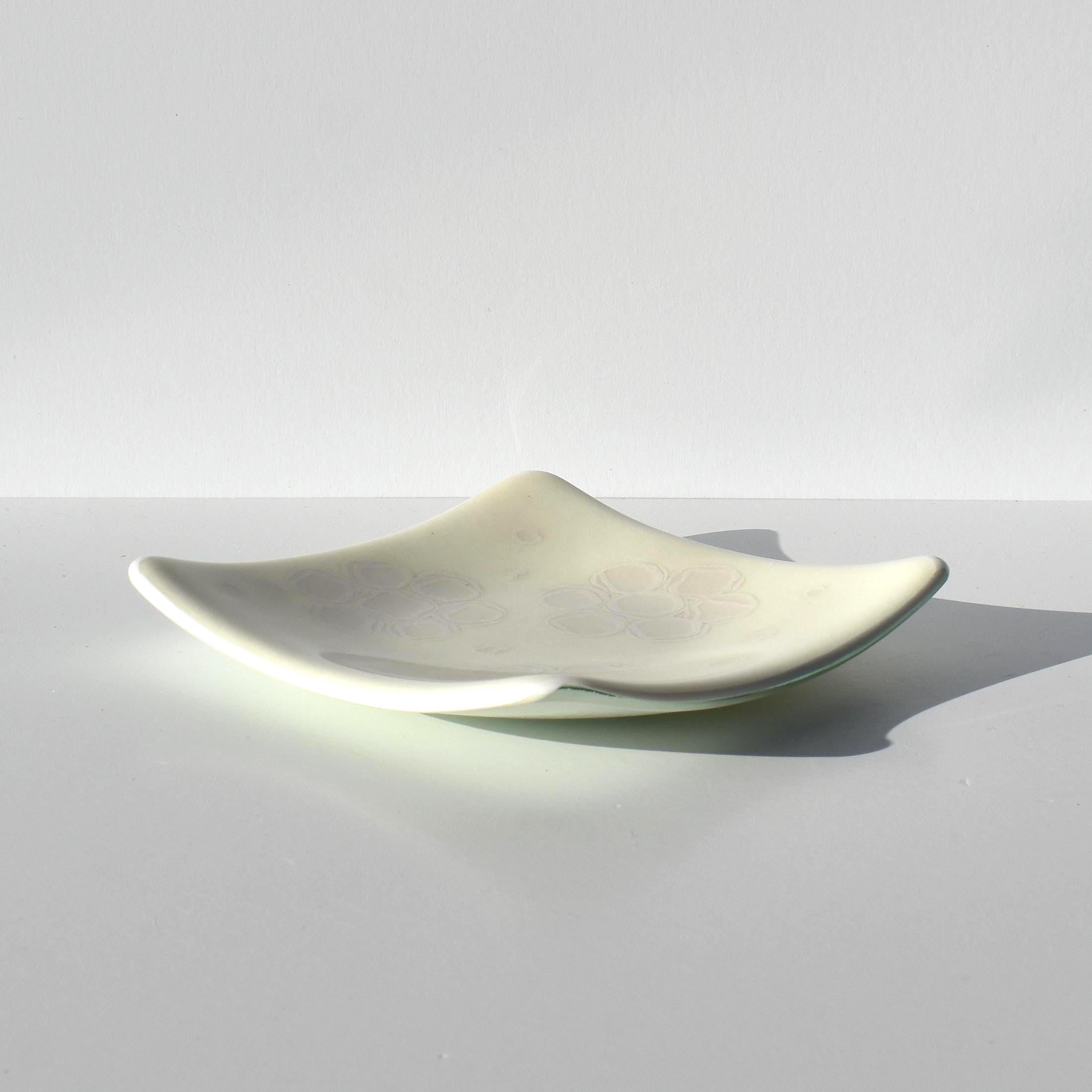 A square glass dish with curved, upturned edges and a smooth, off-white surface. The center of the dish features a subtle, faint floral pattern in a light gray tone, integrated into the translucent material of the glass.