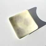 A square, off-white glass dish with rounded corners and a smooth, glossy finish. The surface features three distinct, light-colored floral patterns, each composed of six petal-like shapes arranged in a circular, cluster-like formation.