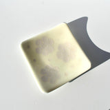 A square, off-white glass dish with rounded corners and a smooth, glossy finish. The surface features three distinct, light-colored floral patterns, each composed of six petal-like shapes arranged in a circular, cluster-like formation.
