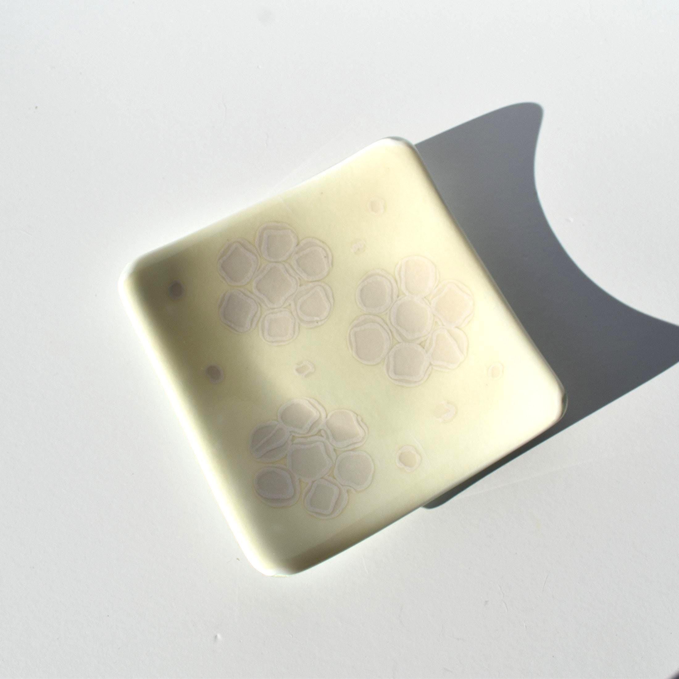 A square, off-white glass dish with rounded corners and a smooth, glossy finish. The surface features three distinct, light-colored floral patterns, each composed of six petal-like shapes arranged in a circular, cluster-like formation.