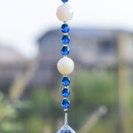 Two large round milky quartz stone beads are strung on wire and hanging vertically, flanked by deep aqua glass crystal beads, anchored by large glass crystal prism; against blurred suburban background