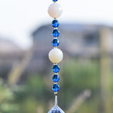 Two large round milky quartz stone beads are strung on wire and hanging vertically, flanked by deep aqua glass crystal beads, anchored by large glass crystal prism; against blurred suburban background