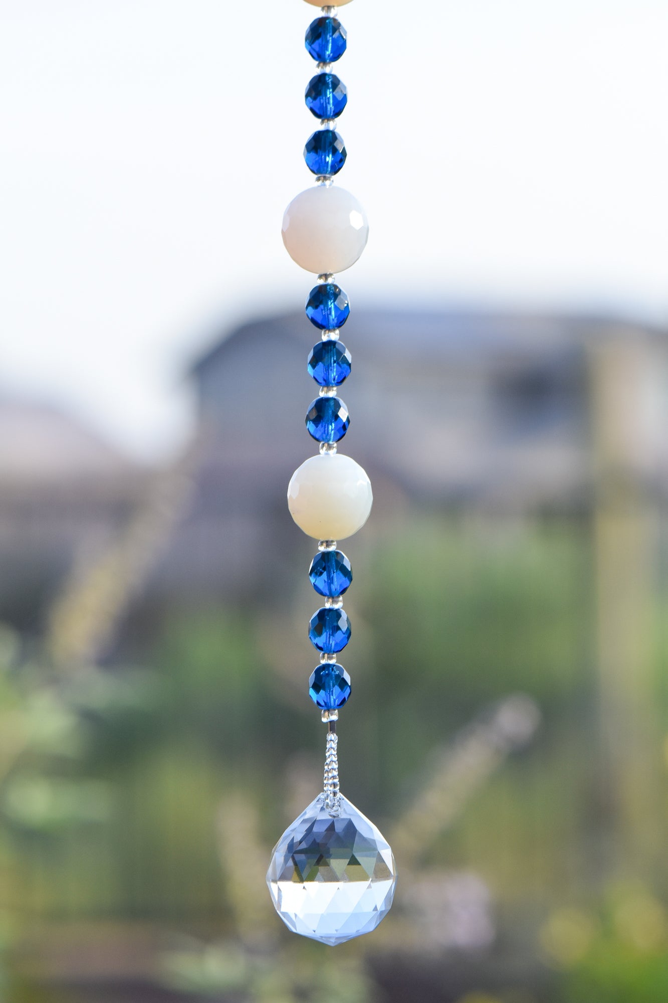 Two large round milky quartz stone beads are strung on wire and hanging vertically, flanked by deep aqua glass crystal beads, anchored by large glass crystal prism; against blurred suburban background
