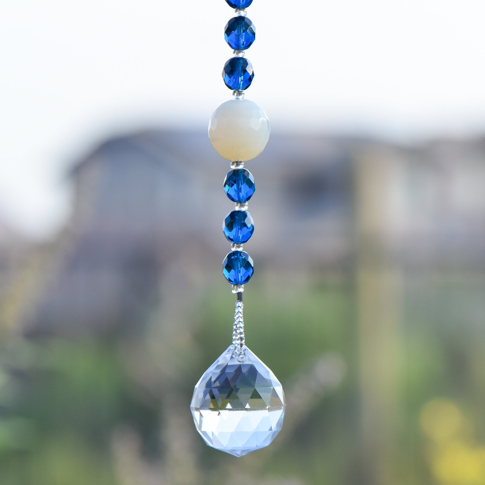 A single large round milky quartz stone beads is strung on wire and hanging vertically, flanked by six deep aqua glass crystal beads, anchored by large glass crystal prism.