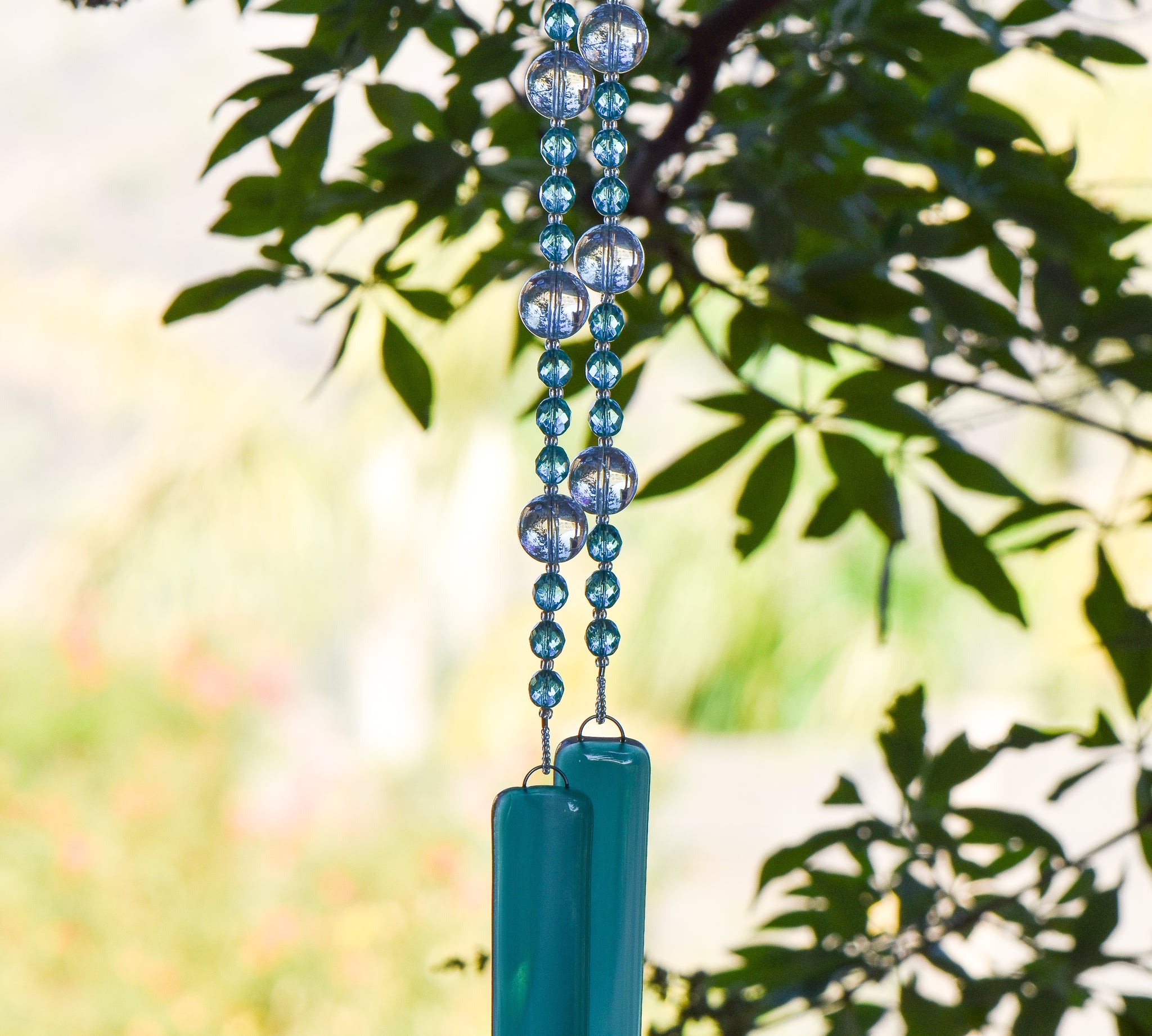 teal green glass beads combined with large reflective glass clear beads, strung on wire and hanging vertically. Anchored by two teal green pieces of fused glass. Hanging from tree.