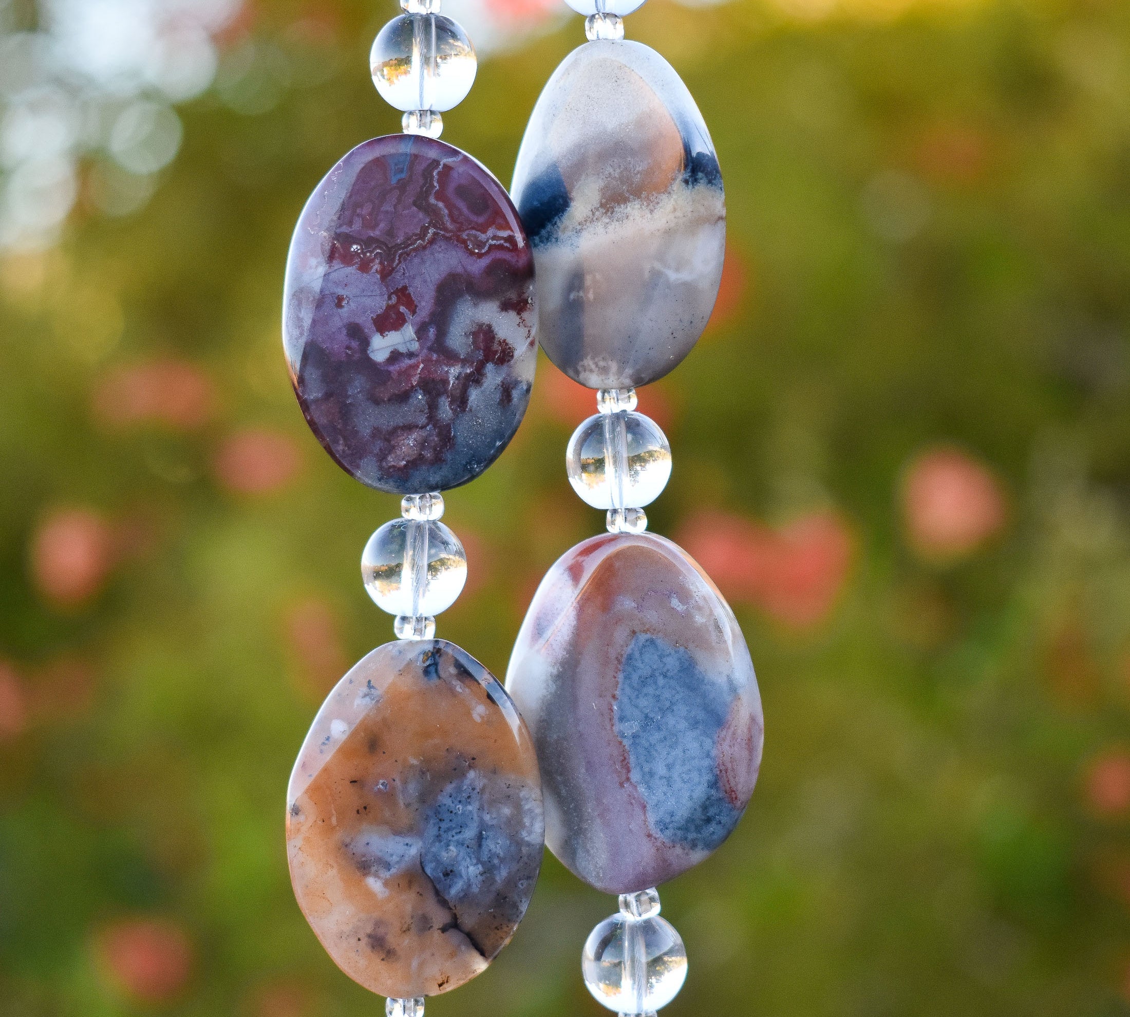 Large, oval-shaped jasper stone beads strung with smaller clear glass beads, hanging vertically in front of plant with orange flowers.