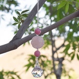 Large carved rose quartz bead flanked by small pink stone beads and anchored by a small crystal prism, all hanging vertically from a tree.