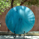 Large, round turquoise blue glass dish with ruffled edges placed in stand in window.