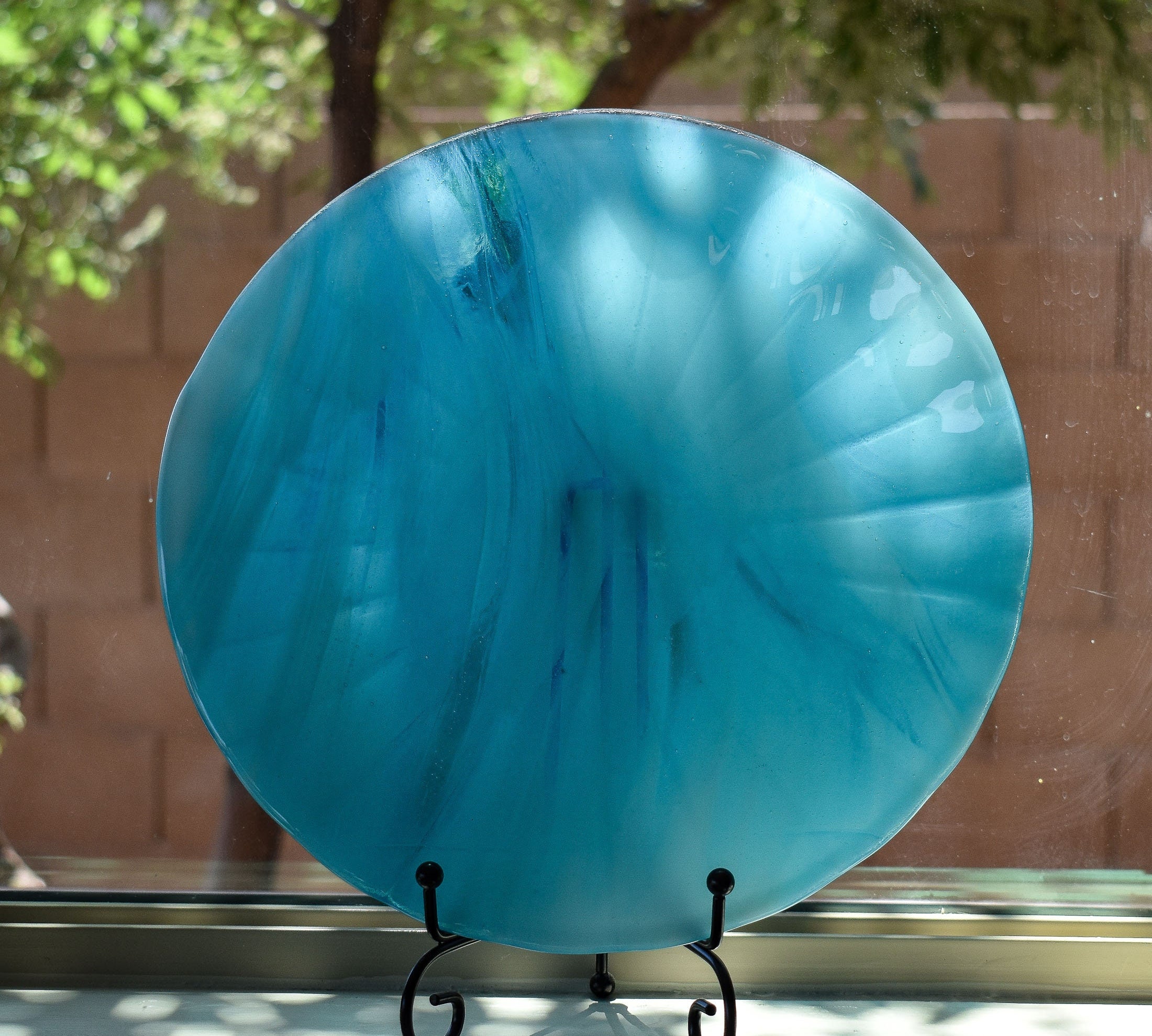 Large, round turquoise blue glass dish with ruffled edges placed in stand in window.