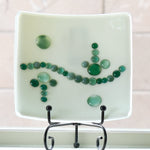Small cream-colored square dish on stand in window. Dish has a wave-like design made with glass dots in various shades of green.