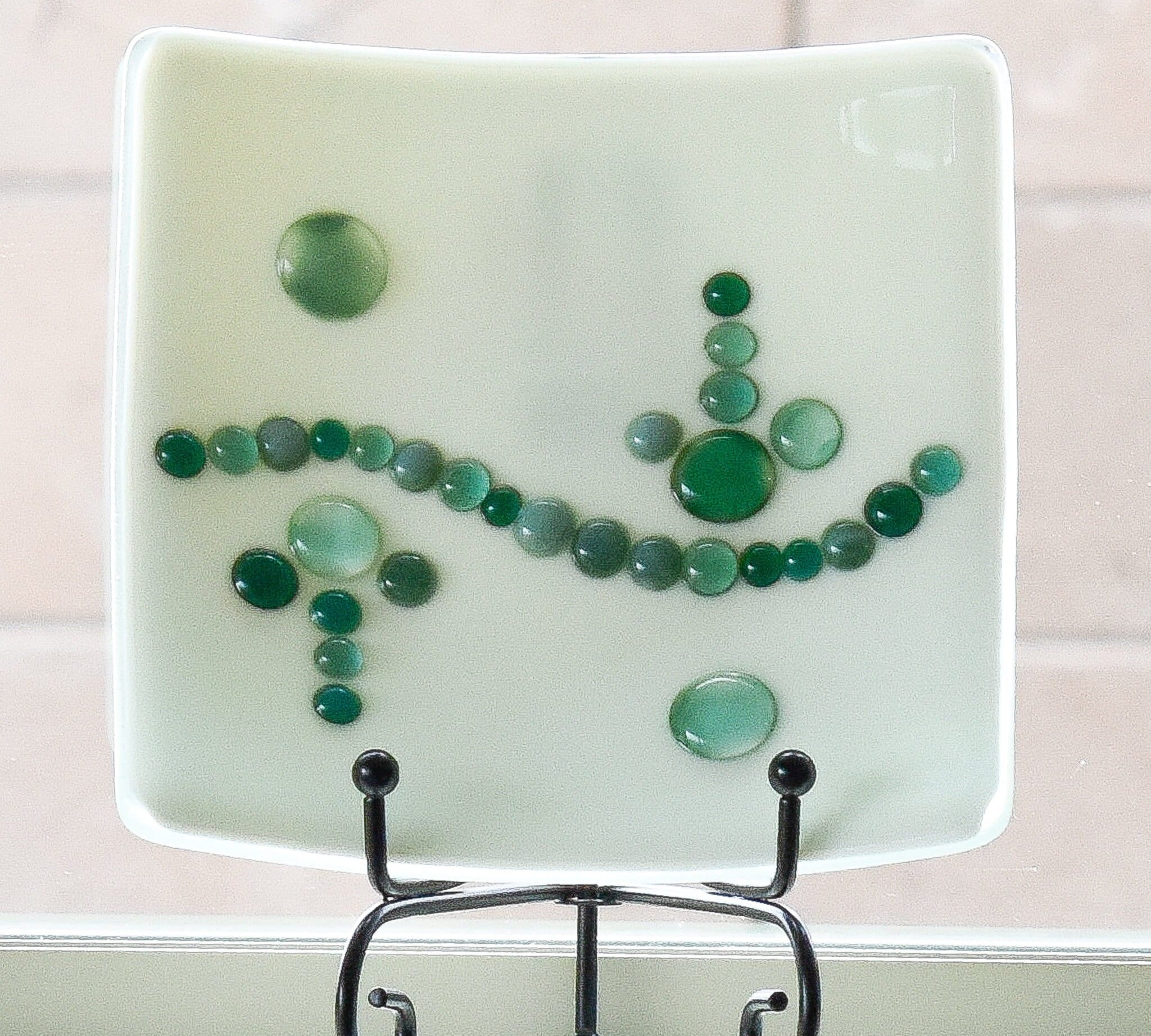 Small cream-colored square dish on stand in window. Dish has a wave-like design made with glass dots in various shades of green.