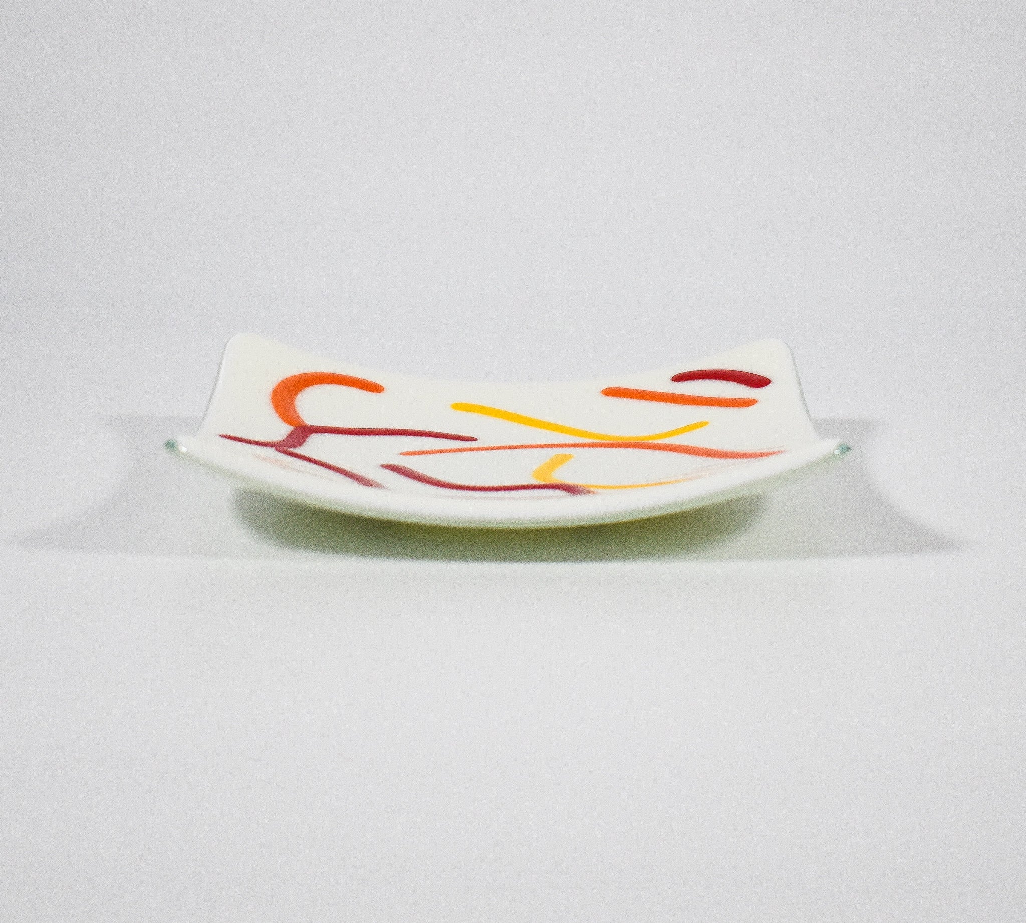 Small, square fused glass tray is cream with orange, red and yellow curve design.