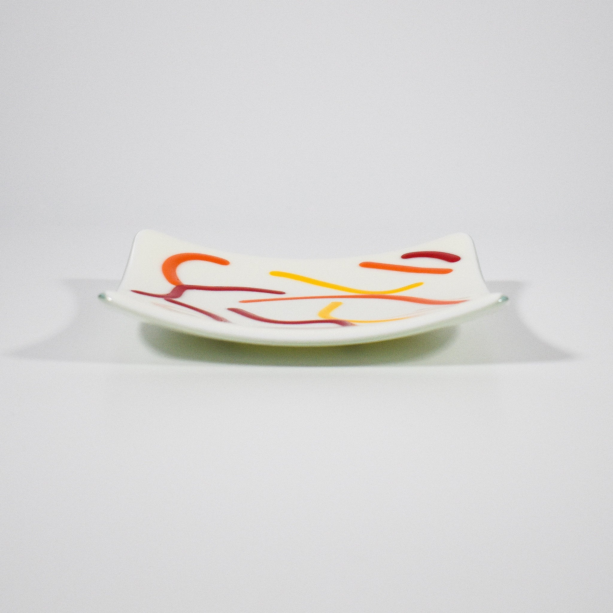 Small, square fused glass tray is cream with orange, red and yellow curve design.
