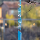 Large blue and white rectangular beads are strung with smaller turquoise blue glass beads and large clear glass beads, all anchored by two pieces of blue fused glass, hanging from tree