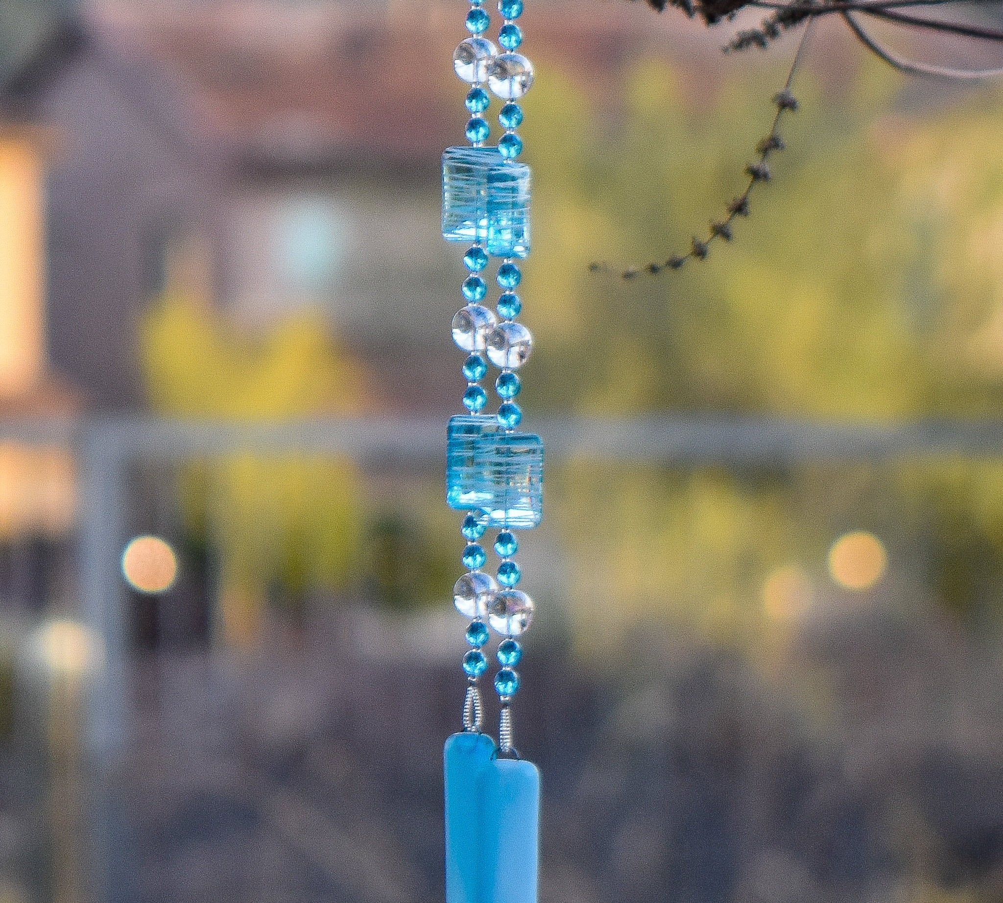 Large blue and white rectangular beads are strung with smaller turquoise blue glass beads and large clear glass beads, all anchored by two pieces of blue fused glass, hanging from tree