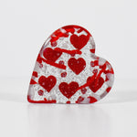 Heart-shaped glass paperweight standing on edge. Paperweight has six red hearts fused into the glass.