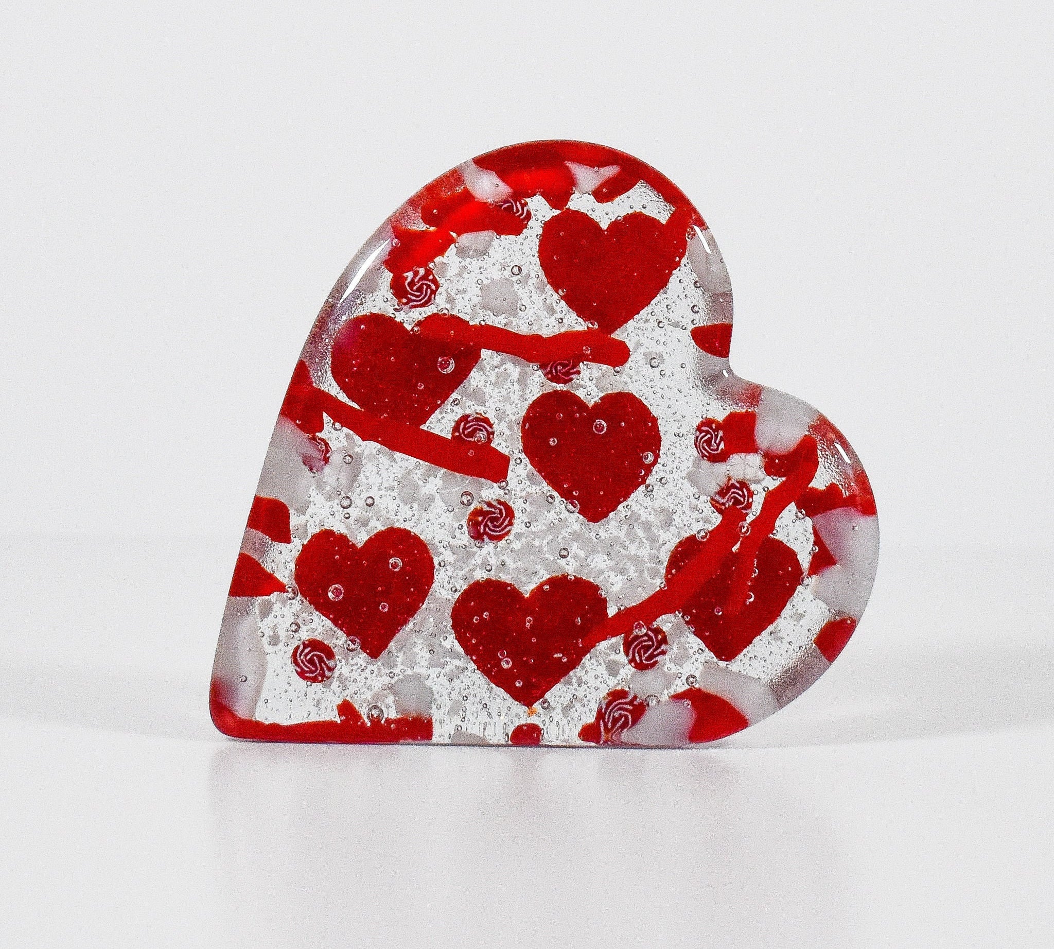 Heart-shaped glass paperweight standing on edge. Paperweight has six red hearts fused into the glass.